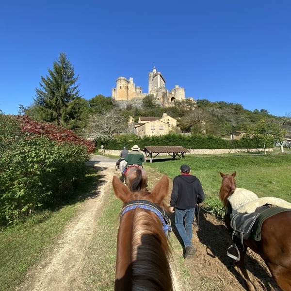 Des cavaliers randonneurs arrivent au château de Bonaguil dans le Lot et Garonne . Les cavaliers marchent sur le chemin  de terrain, la végétation est vert, on aperçoit le lavoir et une maison en pierre, le château trône sur un mamelon avec un ciel bleu sans nuage et une journée ensoleillée